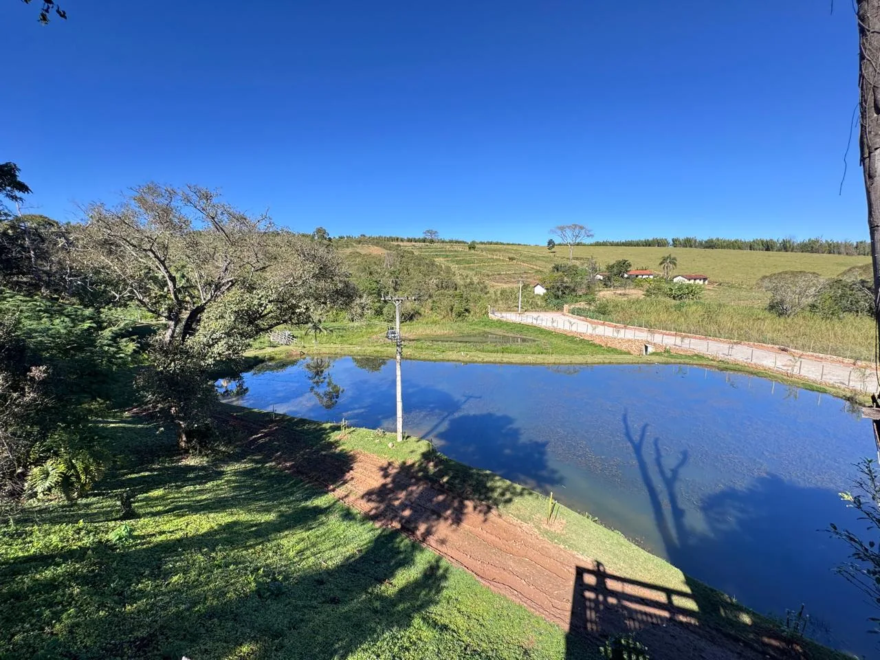 Lago do Residencial Mariah, com árvores, margem verde e paisagem ao fundo